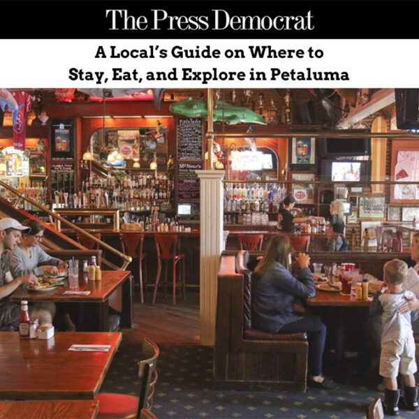 The interior of McNear's Saloon & Dining Hall with multiple people sitting at booths eating, and the bar and employees in the background, Banner at the top features the Press Democrat logo and text overlay that reads, "A Local's Guide on Where to Stay, Eat, and Explore in Petaluma".