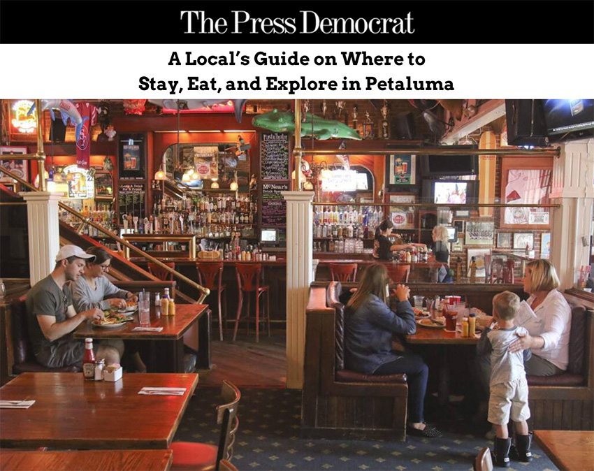 The interior of McNear's Saloon & Dining Hall with multiple people sitting at booths eating, and the bar and employees in the background, Banner at the top features the Press Democrat logo and text overlay that reads, "A Local's Guide on Where to Stay, Eat, and Explore in Petaluma".